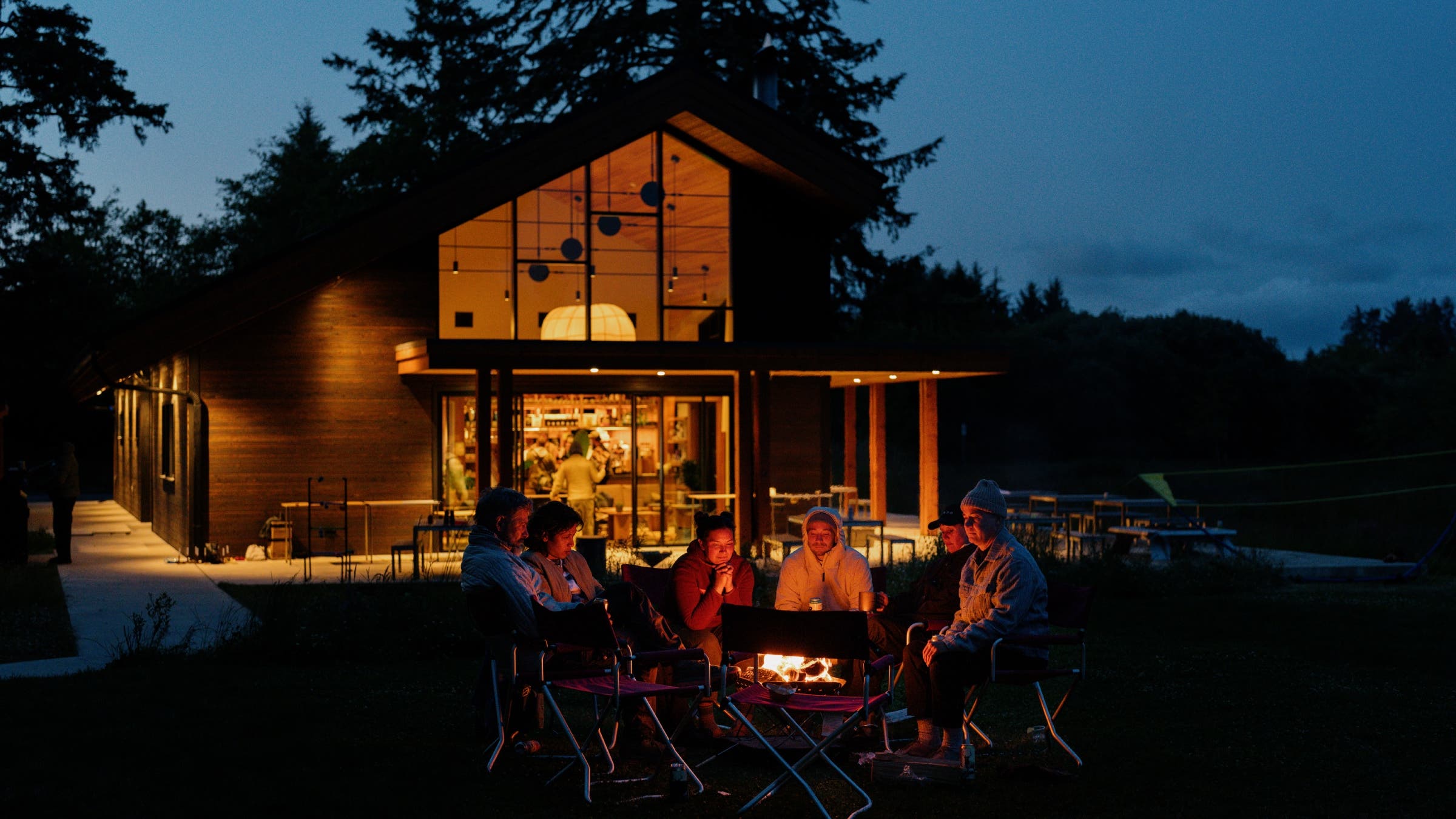 Campers sit around a fire outside of a luxury cabin at dusk.