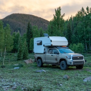 A lifted overlanding truck with big truck tires and a truck bed camper parked in a mountain clearing surrounded by pine and aspen trees at sunset.