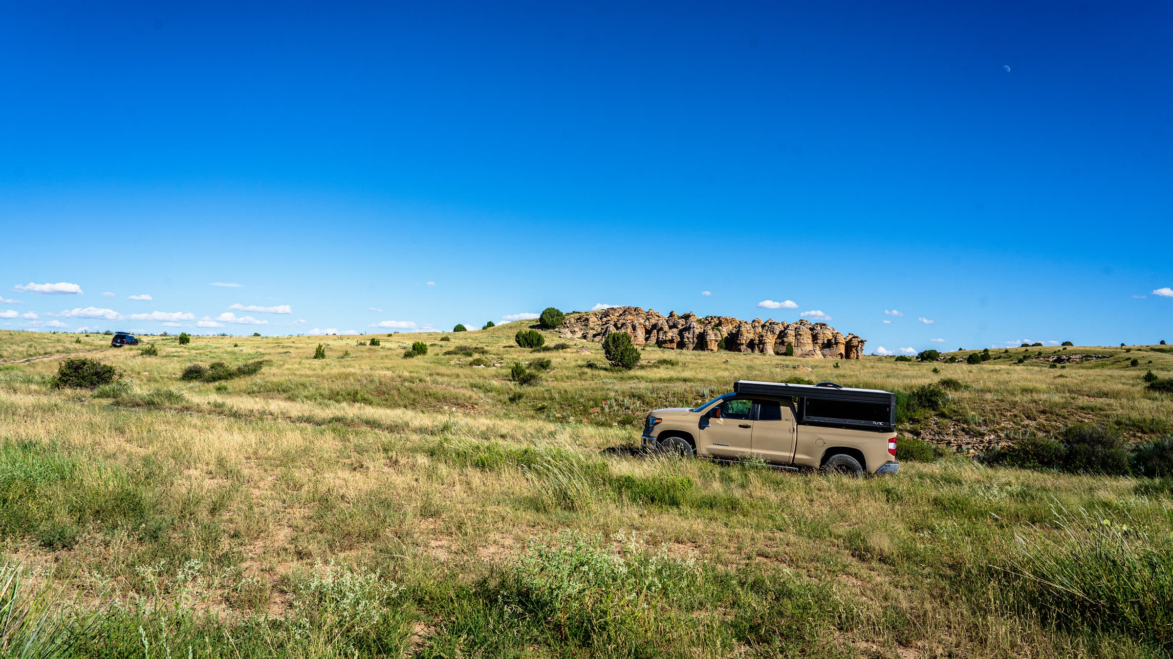 An overland pickup truck with big truck tires and a rooftop camper driving through an open grassland prairie with sandstone rock formations and a vivid blue sky in the background.