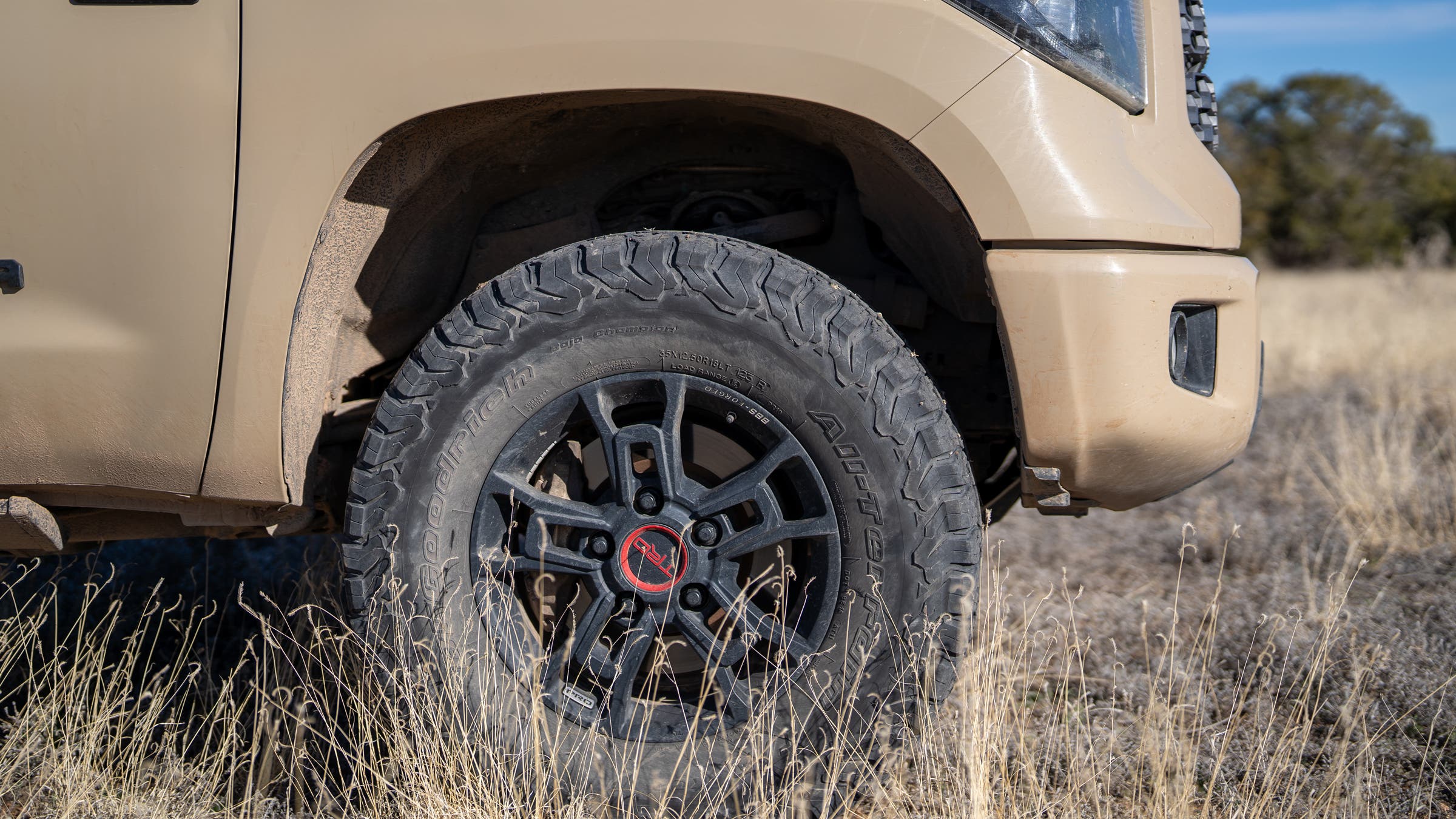 Close-up of big truck tires on a TRD Pro wheel, all-terrain tread pattern mounted on a matte black alloy rim, parked in dry grass off-road terrain.