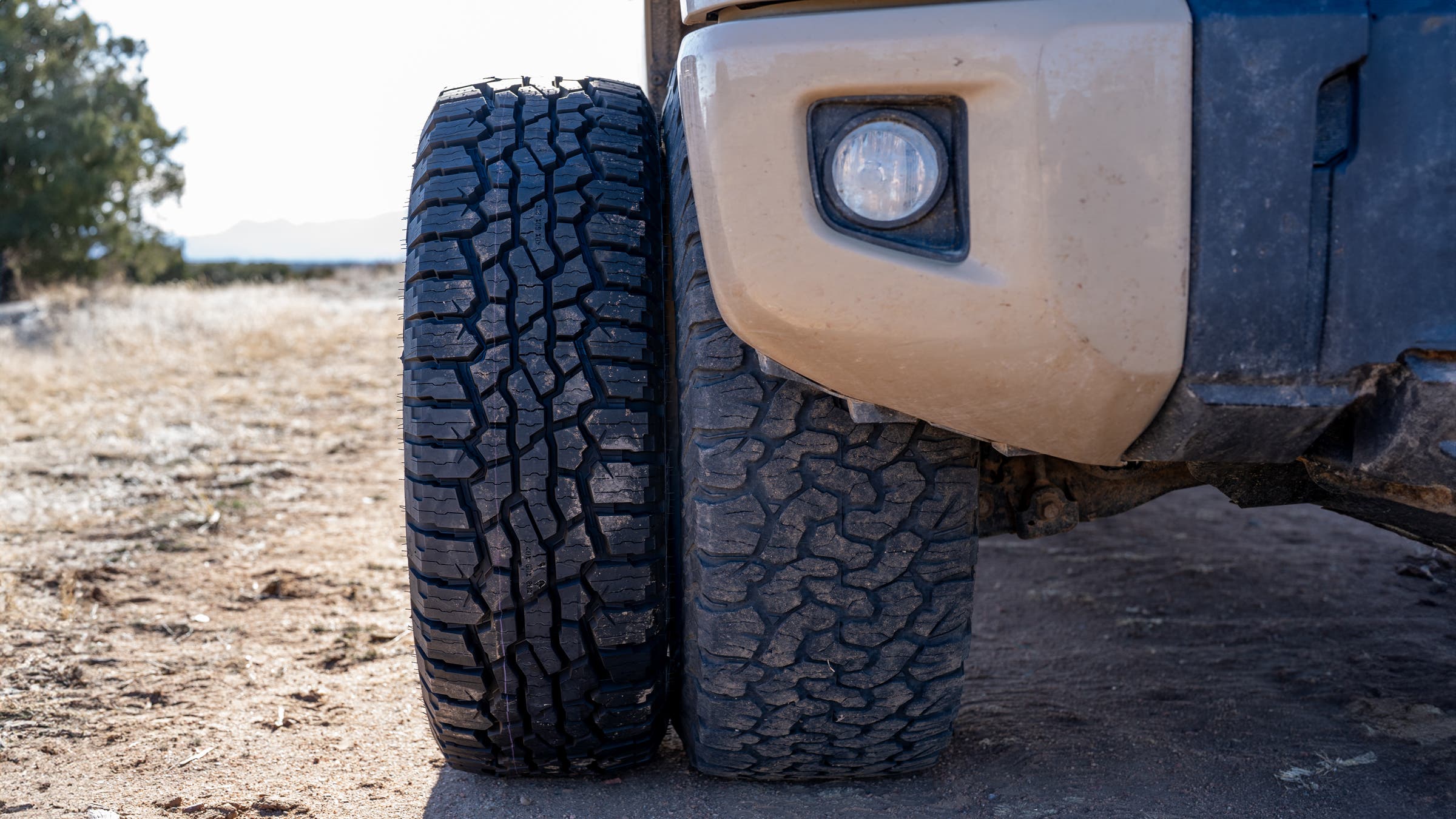 Side-by-side size comparison of big truck tires next to a smaller all-terrain tire leaning against an off-road truck bumper in a dry desert landscape.