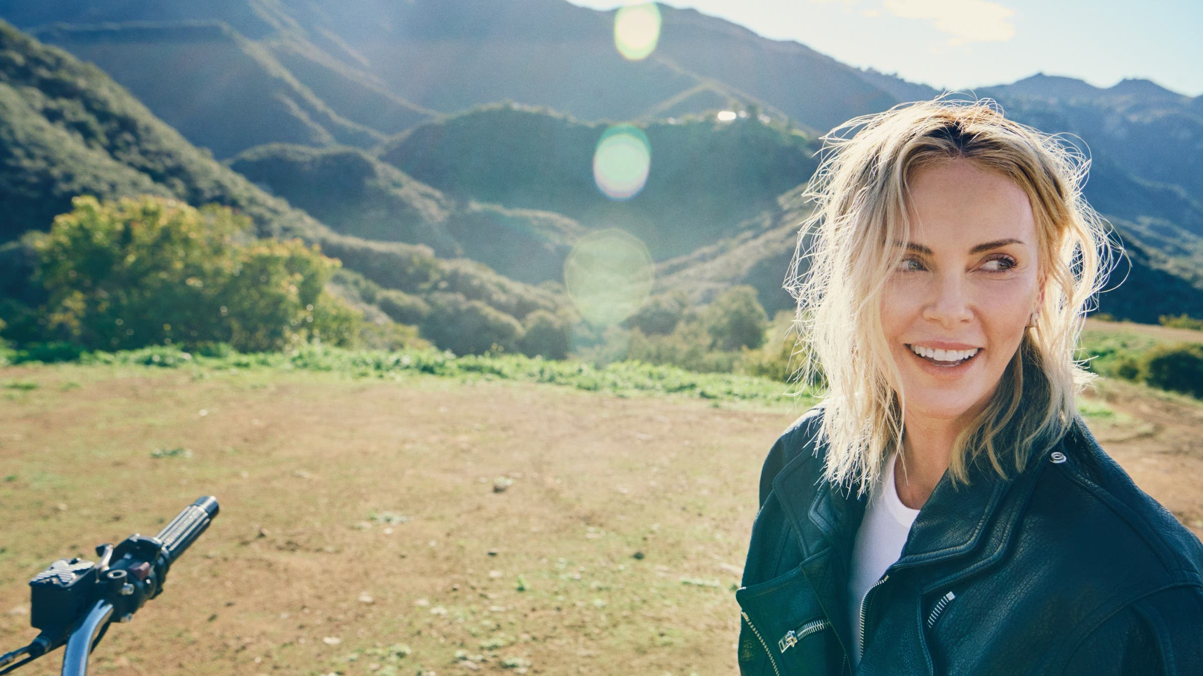Charlize Theron smiling on a sunny day in the mountains.