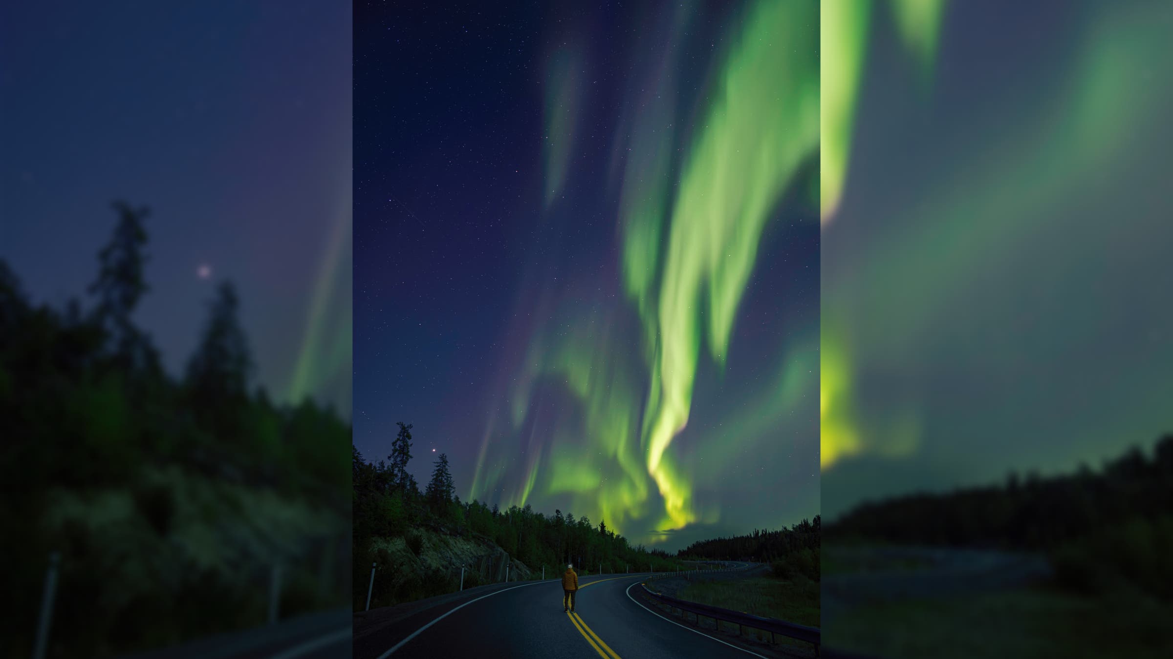 A person stands on an empty road underneath green ribbons of aurora.