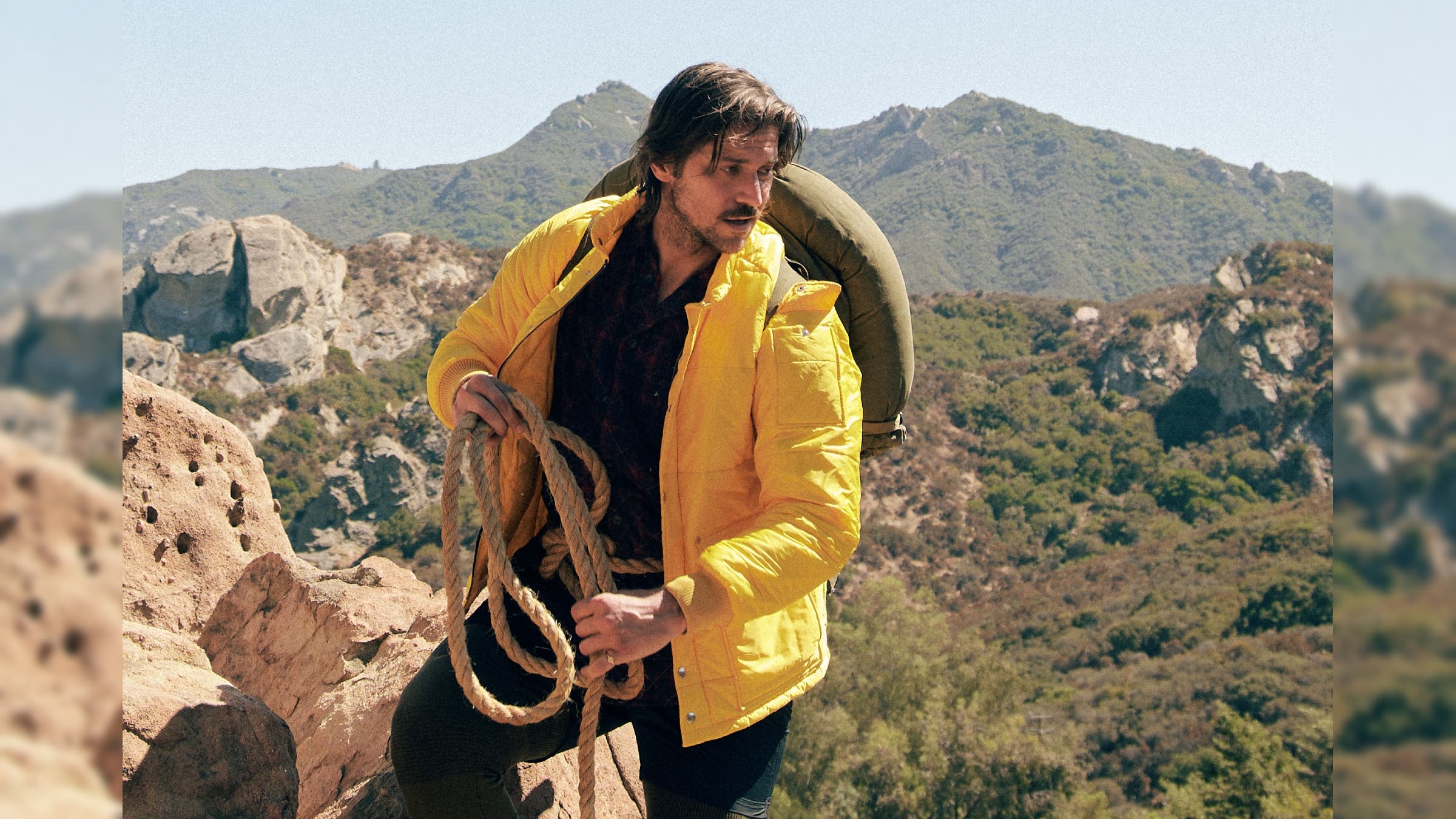 Lucas Bravo in the mountains, holding a rope.