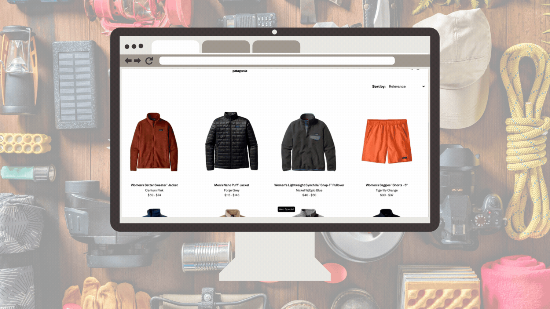 A desktop computer screen displays the Patagonia Worn Wear website, featuring a grid of various used outdoor gear such as puffer jackets, fleece pullovers, and hiking shorts. The monitor is set against a background of organized camping equipment, including ropes, a headlamp, and a water bottle.