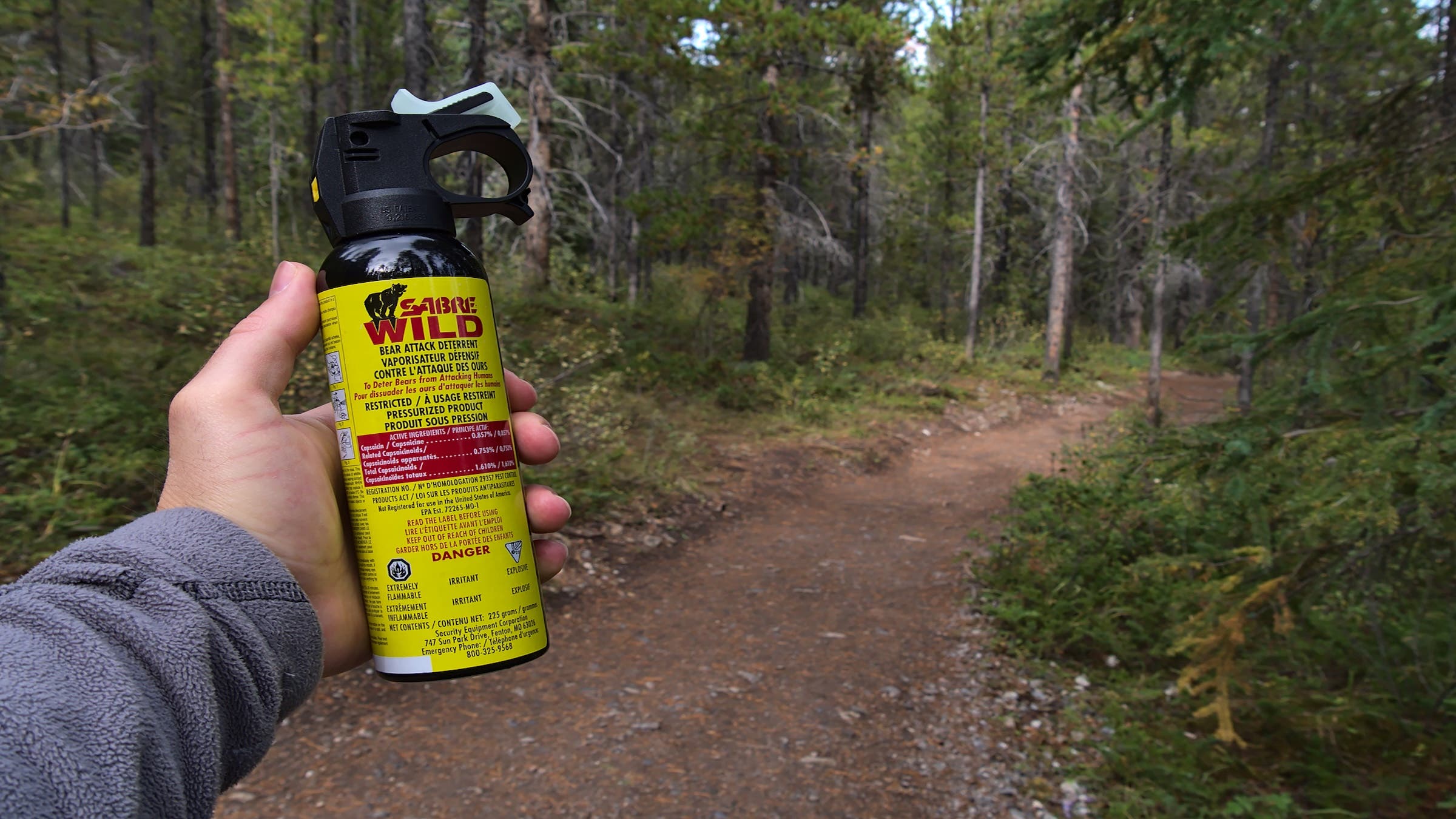 Hiker holding bear spray (Sabre Wild brand) used as bear attack deterrent in forest near Canmore, Canada with hiking trail in background.