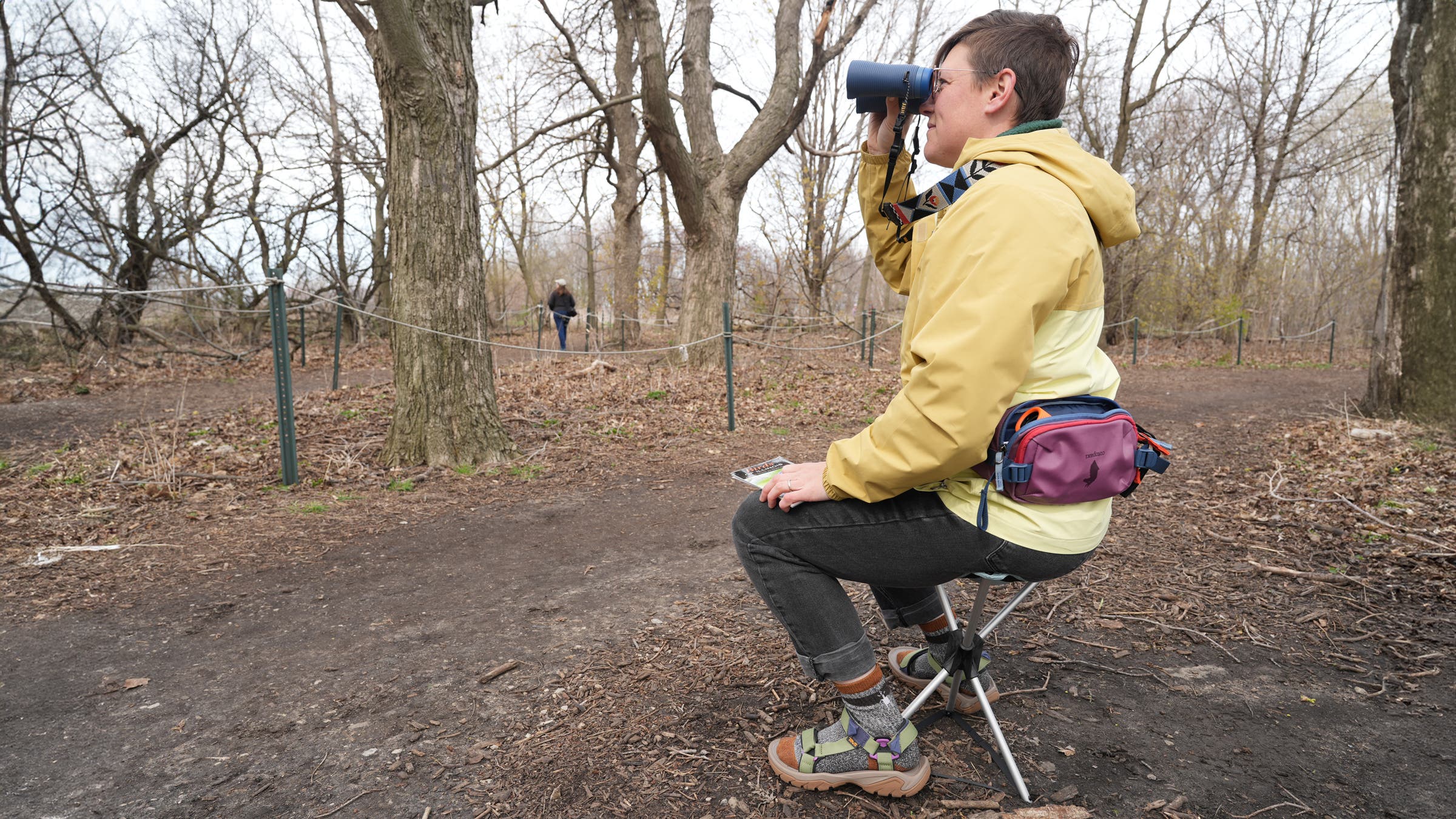 Looking through binoculars while sitting on REI Flexlite Air stool in the woods on a trail while birding, an essential piece of the best birding gear