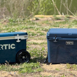 Blue RTIC Ultralight Wheeled 52 QT cooler displayed next to blue Yeti Tundra Haul wheeled cooler on grass