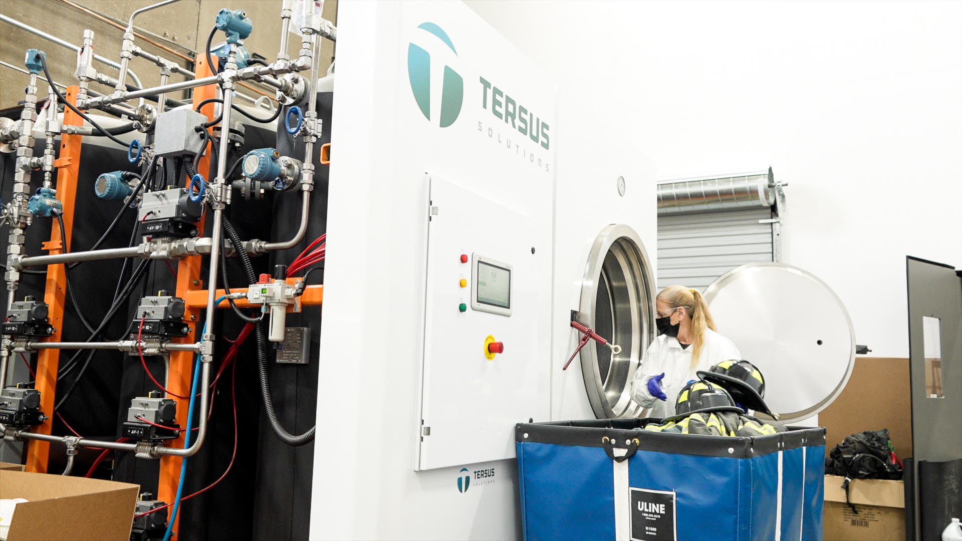 A technician in a white lab coat, gloves, and face mask loads firefighter gear—including a helmet and protective clothing—into a large industrial cleaning machine branded with the Tersus Solutions logo. A complex array of pipes, valves, sensors, and wiring is visible on an orange equipment rack to the left. A blue Uline laundry bin sits in the foreground.