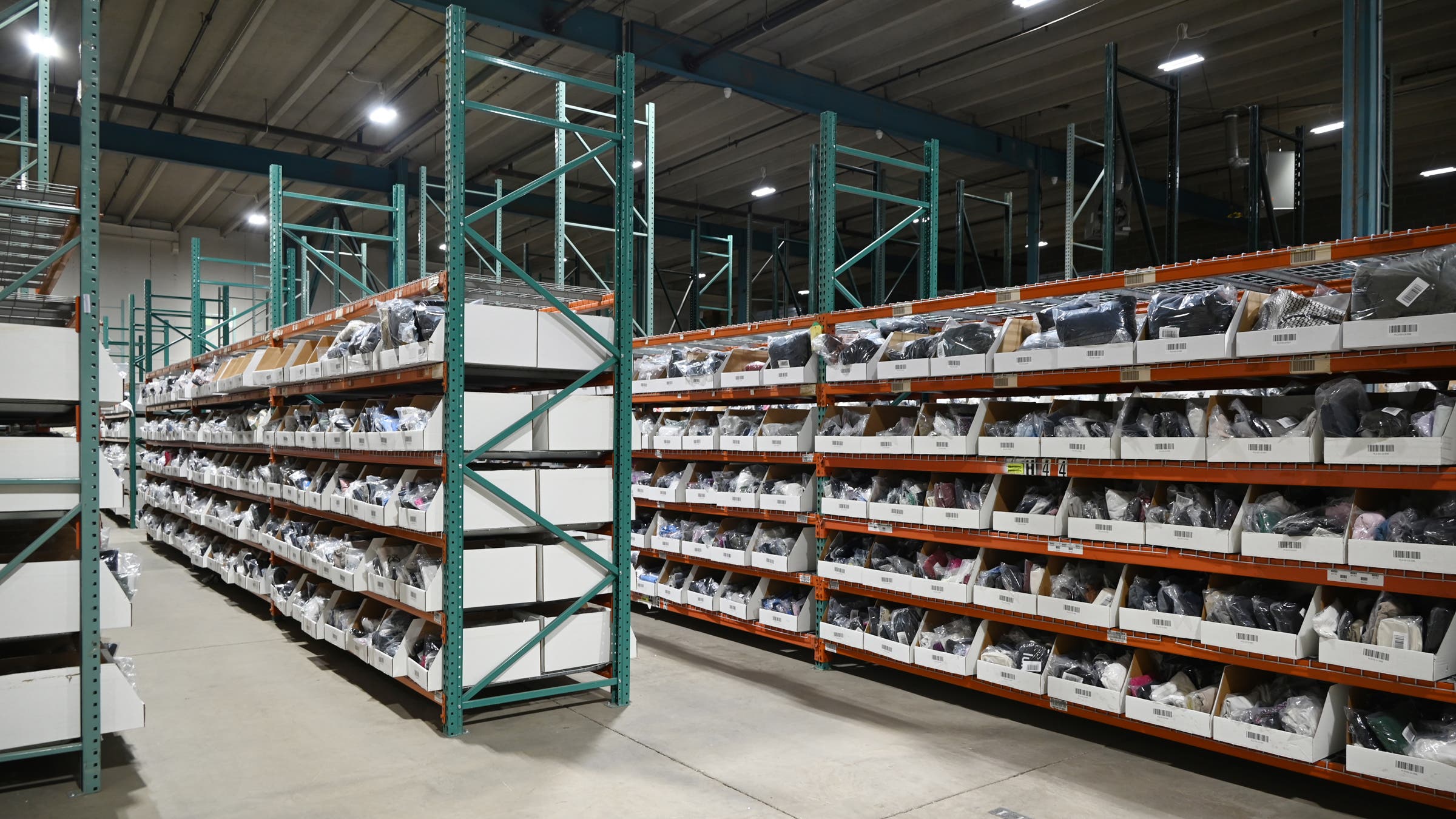 The interior of a large fulfillment warehouse with rows of tall industrial shelving units in orange and teal. The shelves are densely packed with white bin boxes containing individually bagged clothing items. Bright overhead lighting illuminates the concrete floor and high ceiling of the expansive facility.