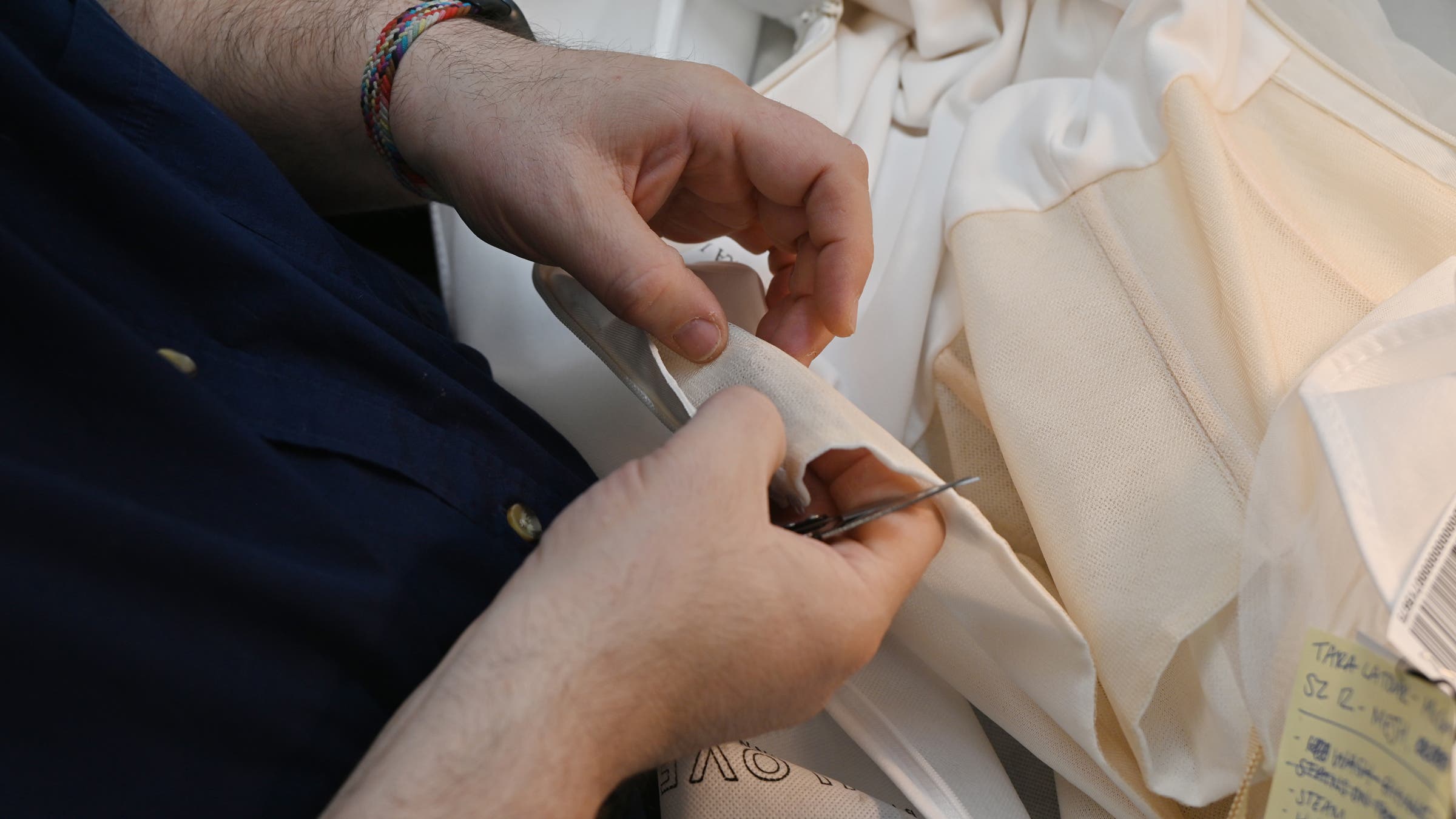 A close-up of a person's hands using small scissors to carefully cut or snip a seam on a cream-colored garment. The person wears a colorful braided bracelet and a navy blue shirt. A handwritten repair tag is visible in the lower right corner, and additional fabric pieces are stacked nearby.