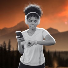 a woman holding her phone and checking her fitness watch. She has headphones over her ears.