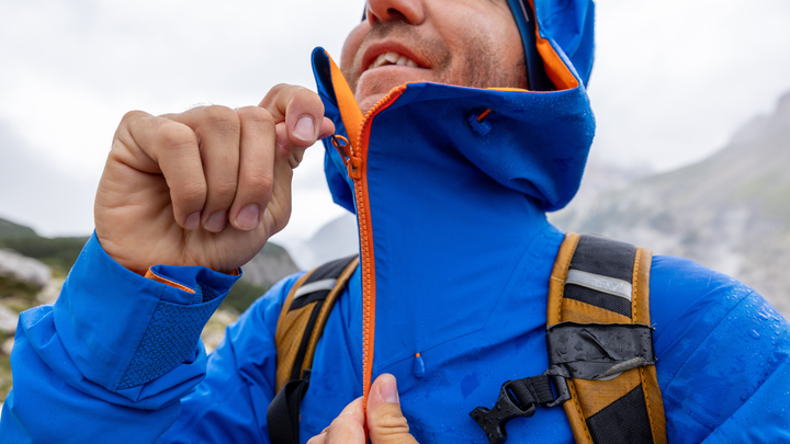 A detailed close-up shot of a person zipping up a waterproof blue rain jacket with bright orange lining, highlighting the tech fabric details often discussed in guides on how to wash a rain jacket.