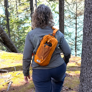Rear view of tester Kelly Bastone hiking through a pine forest wearing the orange REI Co-op Trailmade hydration sling across her back.