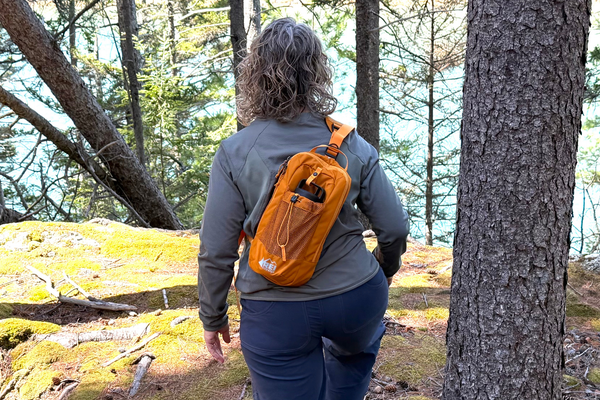 Rear view of tester Kelly Bastone hiking through a pine forest wearing the orange REI Co-op Trailmade hydration sling across her back.
