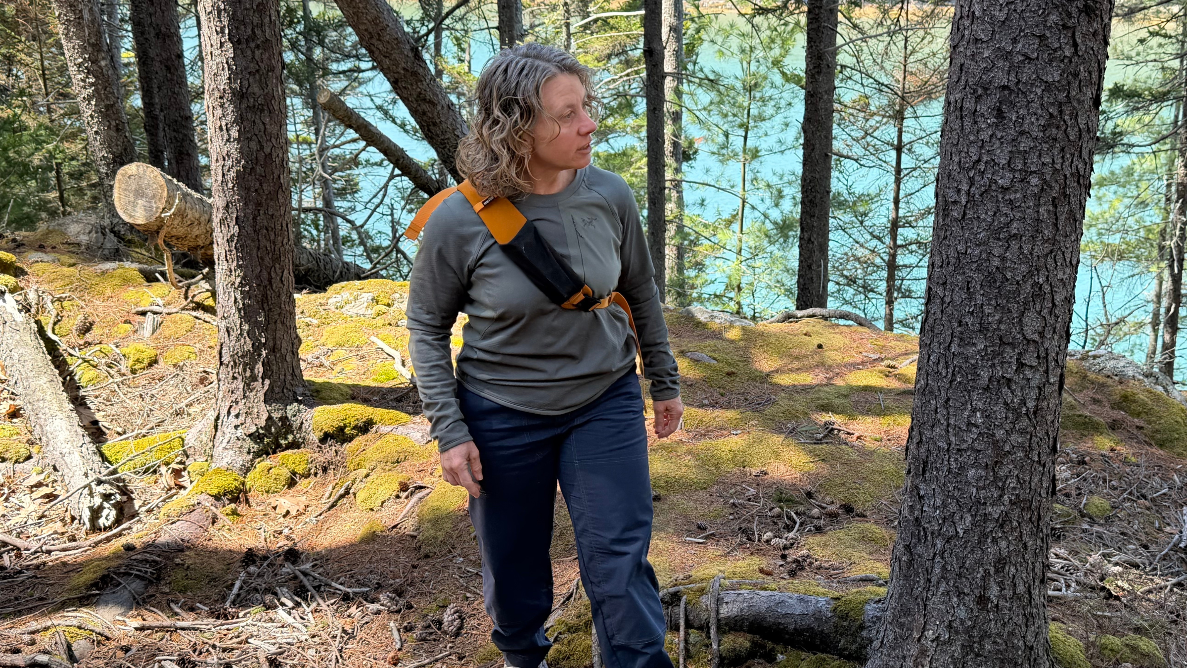 Author Kelly Bastone stands on a mossy forest overlook wearing the REI Co-op Trailmade hydration sling, showing how the padded strap sits across the chest.
