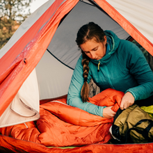 A woman in a blue jacket sits inside a tent, carefully inspecting and fluffing an orange sleeping bag as part of essential sleeping bag care.