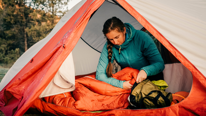 A woman in a blue jacket sits inside a tent, carefully inspecting and fluffing an orange sleeping bag as part of essential sleeping bag care.