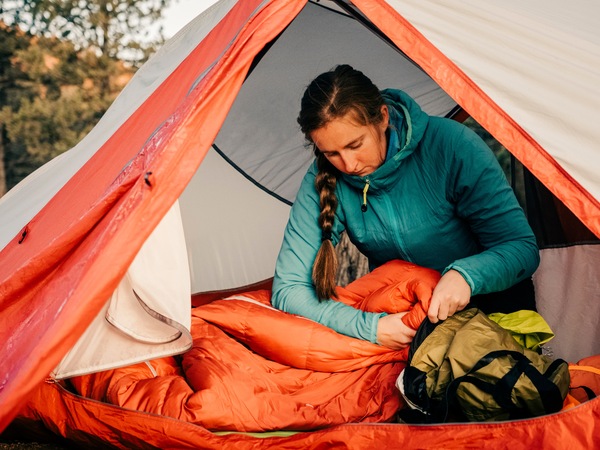 A woman in a blue jacket sits inside a tent, carefully inspecting and fluffing an orange sleeping bag as part of essential sleeping bag care.