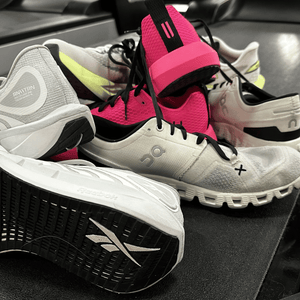 A pile of the best treadmill running shoes, featuring various models from Reebok and On, sits atop a black treadmill belt. The assortment showcases a range of designs, from high-traction outsoles to breathable mesh uppers, specifically selected for indoor performance and stability.