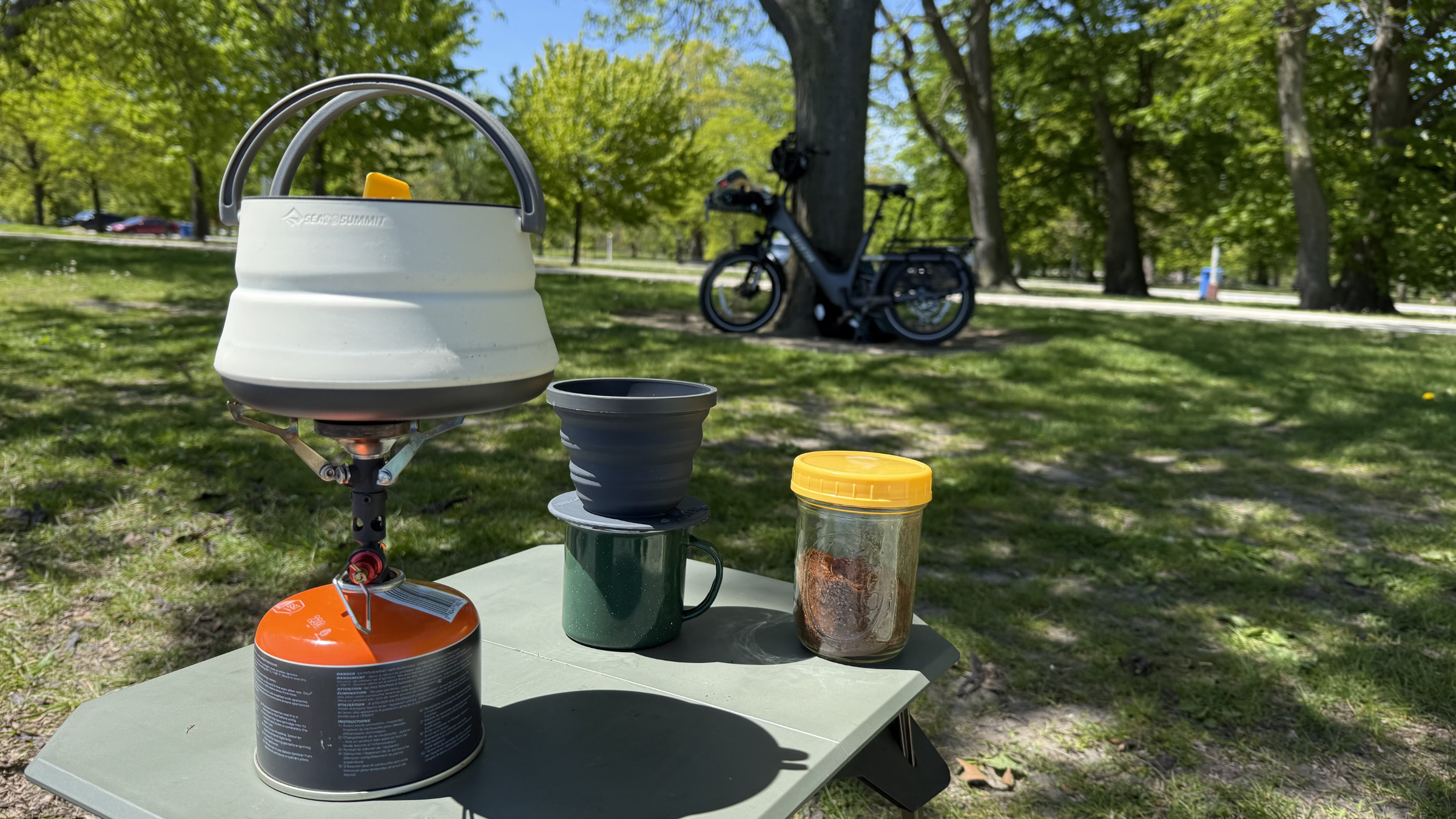 A Sea to Summit collapsible kettle boils water on a portable gas stove next to a pour-over dripper resting on a green enamel mug for a fresh cup of camping coffee.