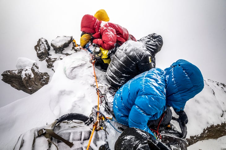 four people are photographed sitting along a snowy ridgeline clipped into their rope