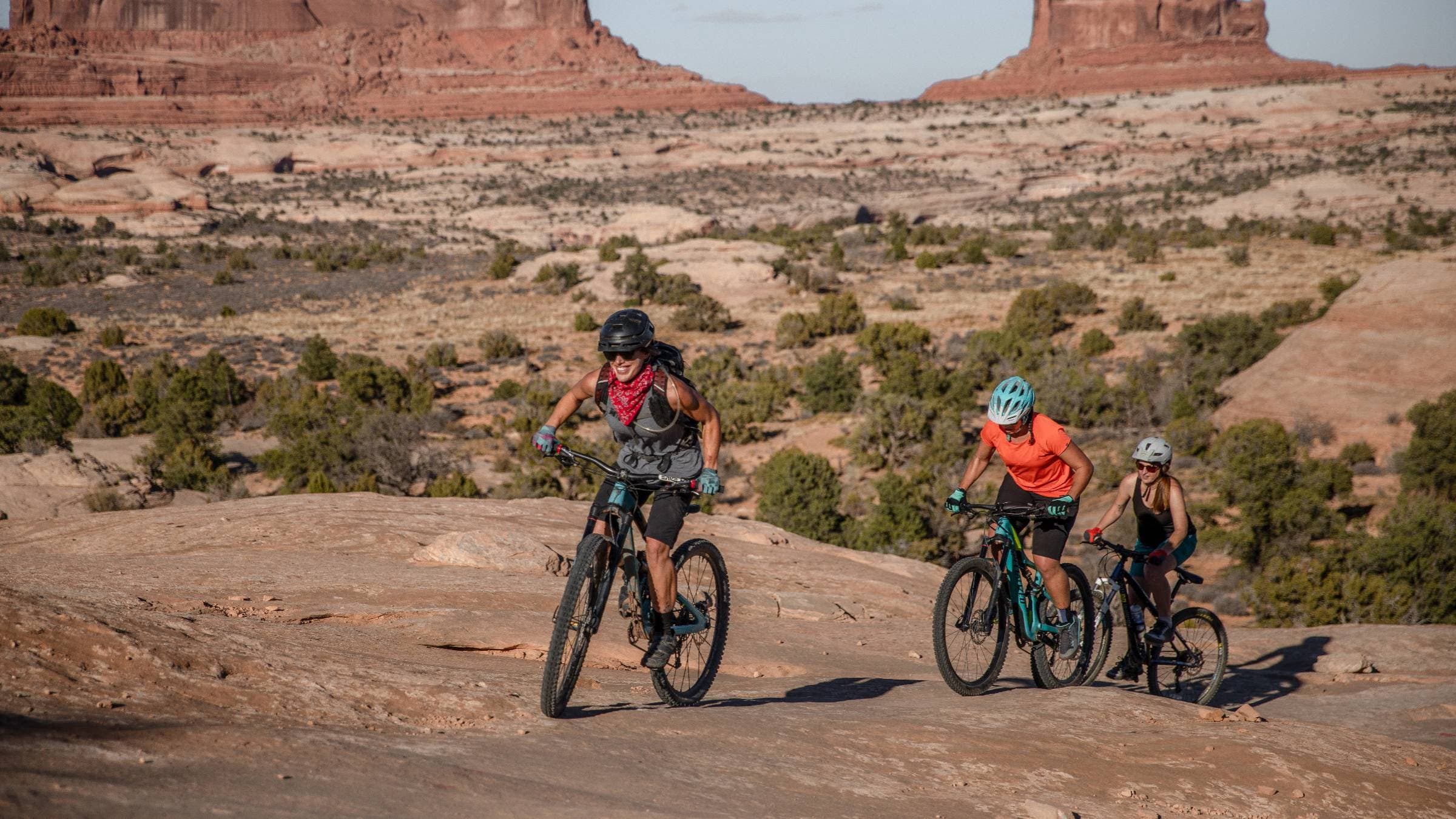 Three women ride mountain bikes uphill over the mountainous terrain of Navajo Rocks Trail in Moab, Utah.