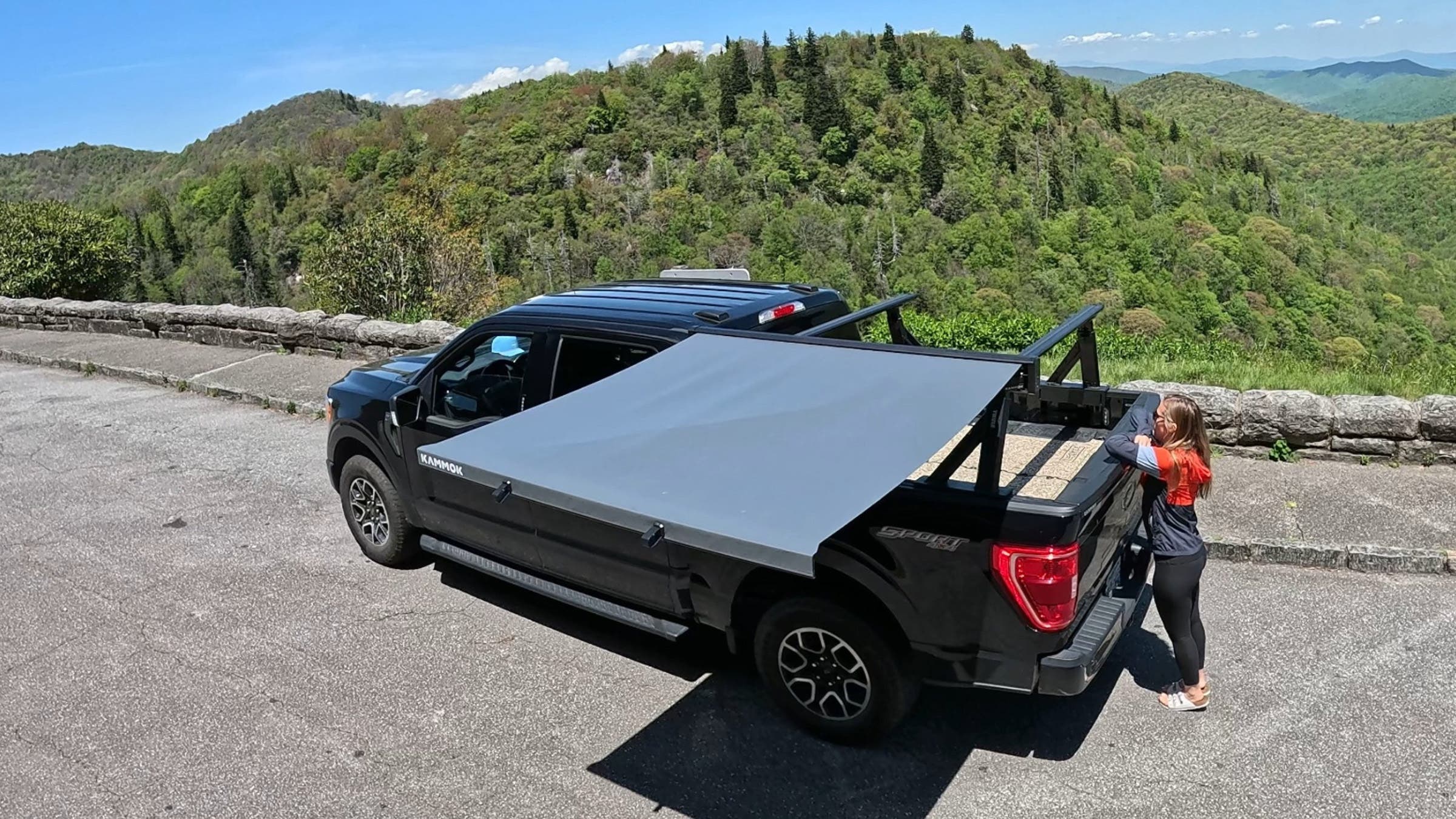 Overhead view of the Kammok Crosswing awning deployed off the side of a black Ford F150, with rolling green mountains visible beyond the parking lot overlook.