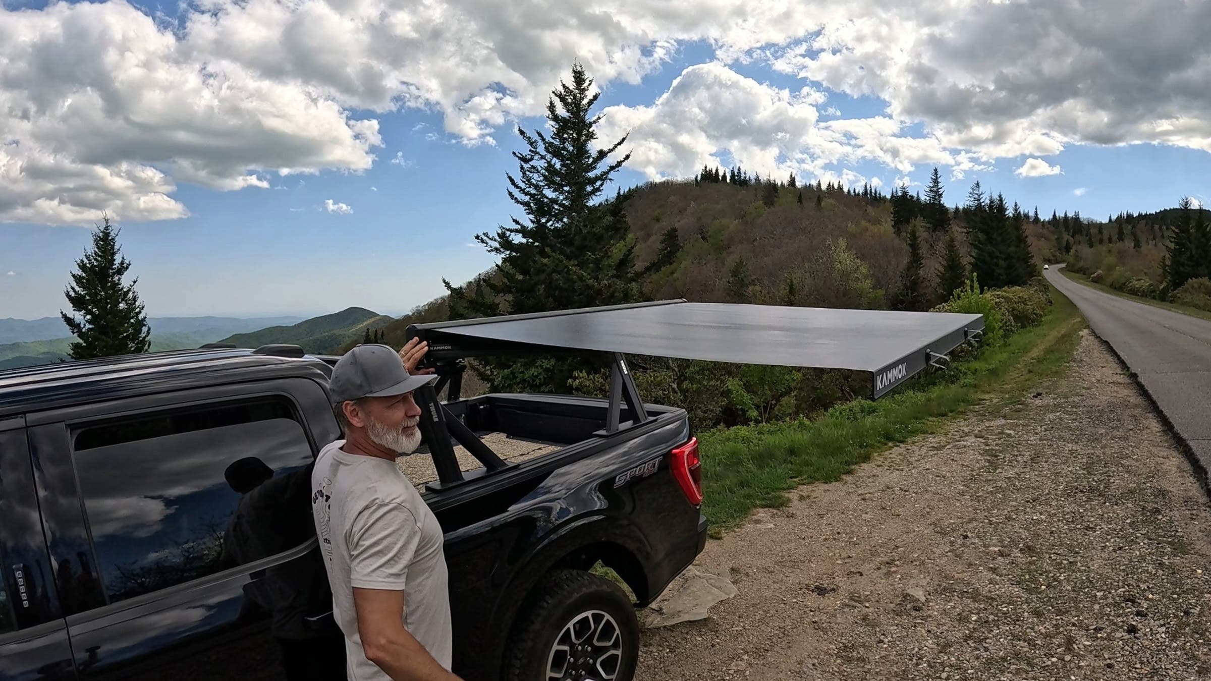 A bearded man in a cap and white t-shirt stands beside a black Ford F150 truck with the Kammok Crosswing awning fully extended.