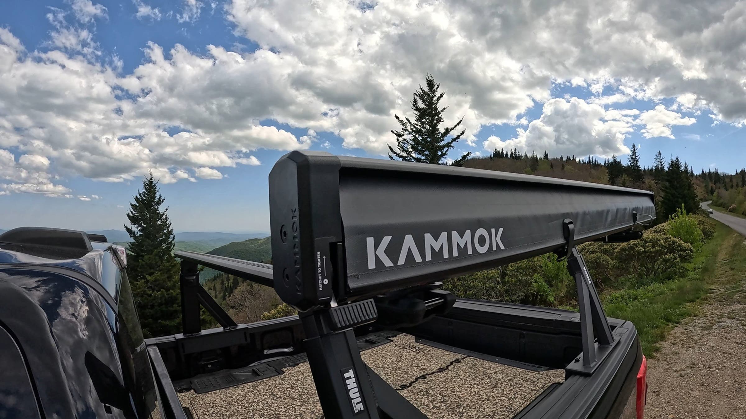 Close-up of the Kammok Crosswing awning housing mounted on a Thule XScape truck rack, showing the ratchet tensioning system and Kammok branding, with mountain scenery in the background.