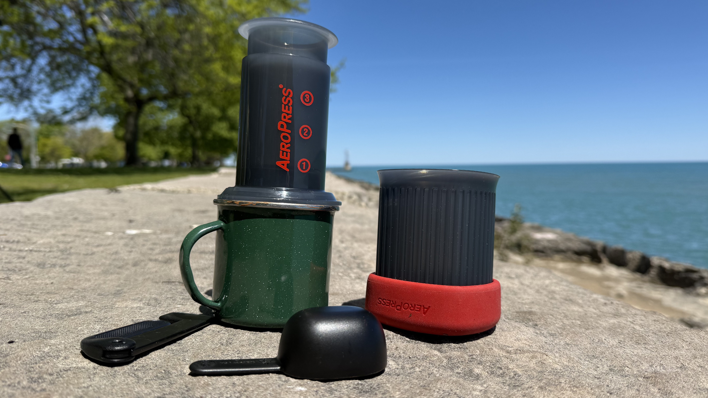 An AeroPress Go travel coffee press sits on top of a green speckled enamelware mug on a concrete seawall by a blue lake, ready to press fresh camping coffee.