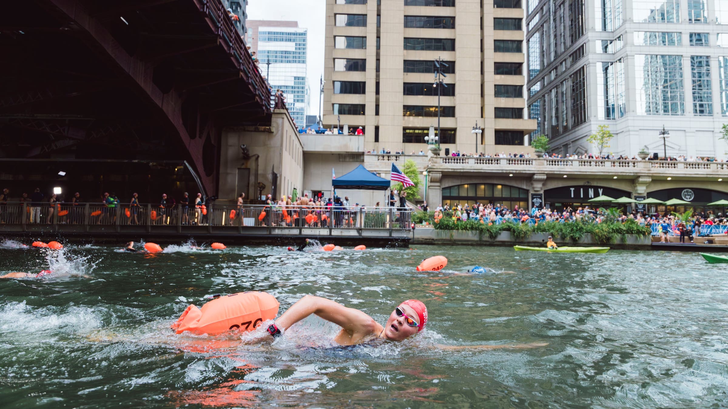 Swimmers racing in a river surrounded by tall buildings.