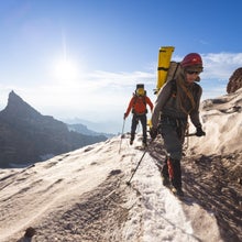 Two men scale a mountain with a prominent mountain peak over their shoulders.