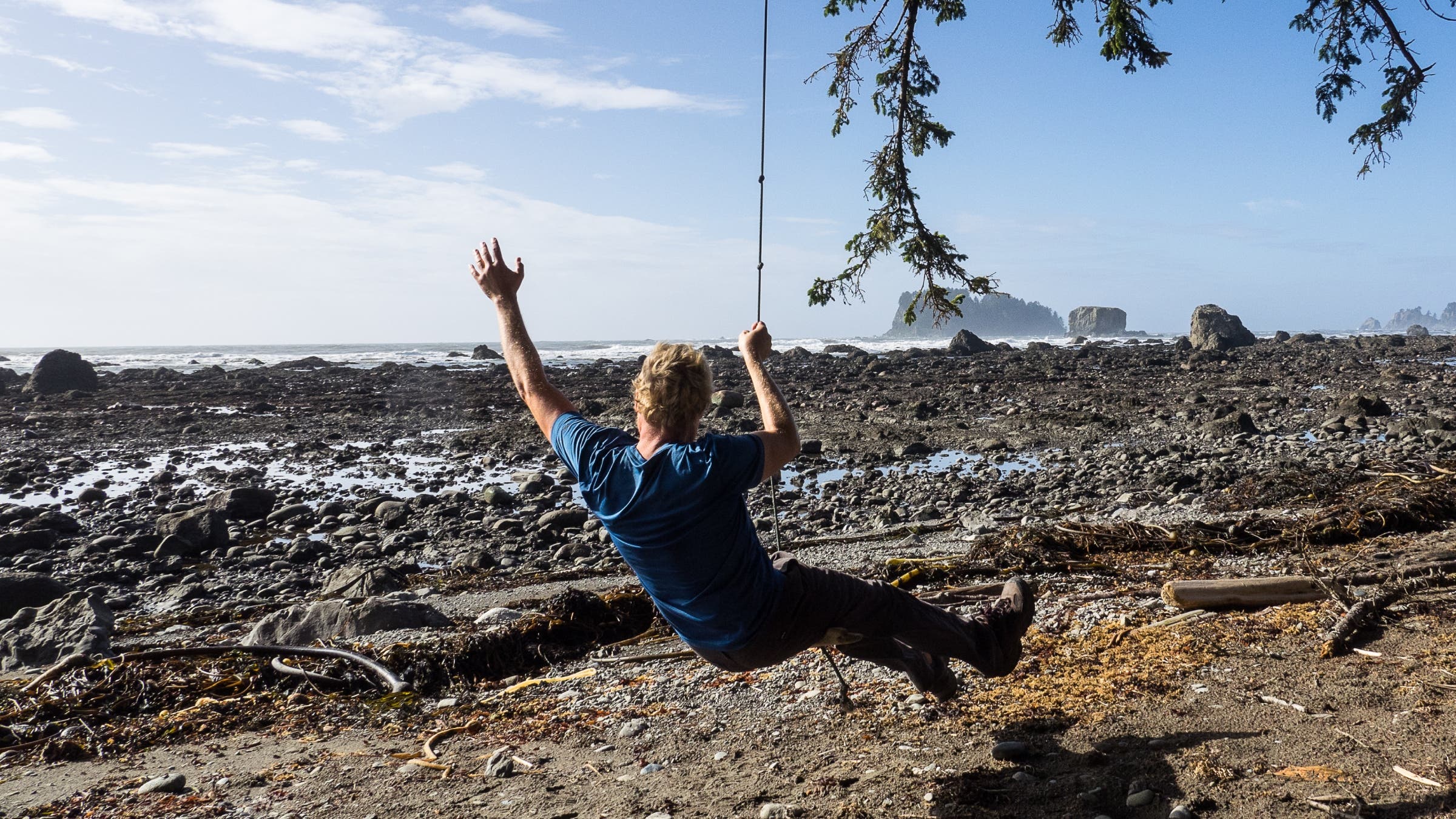 Man swinging from a tree on a wide, rugged beach.