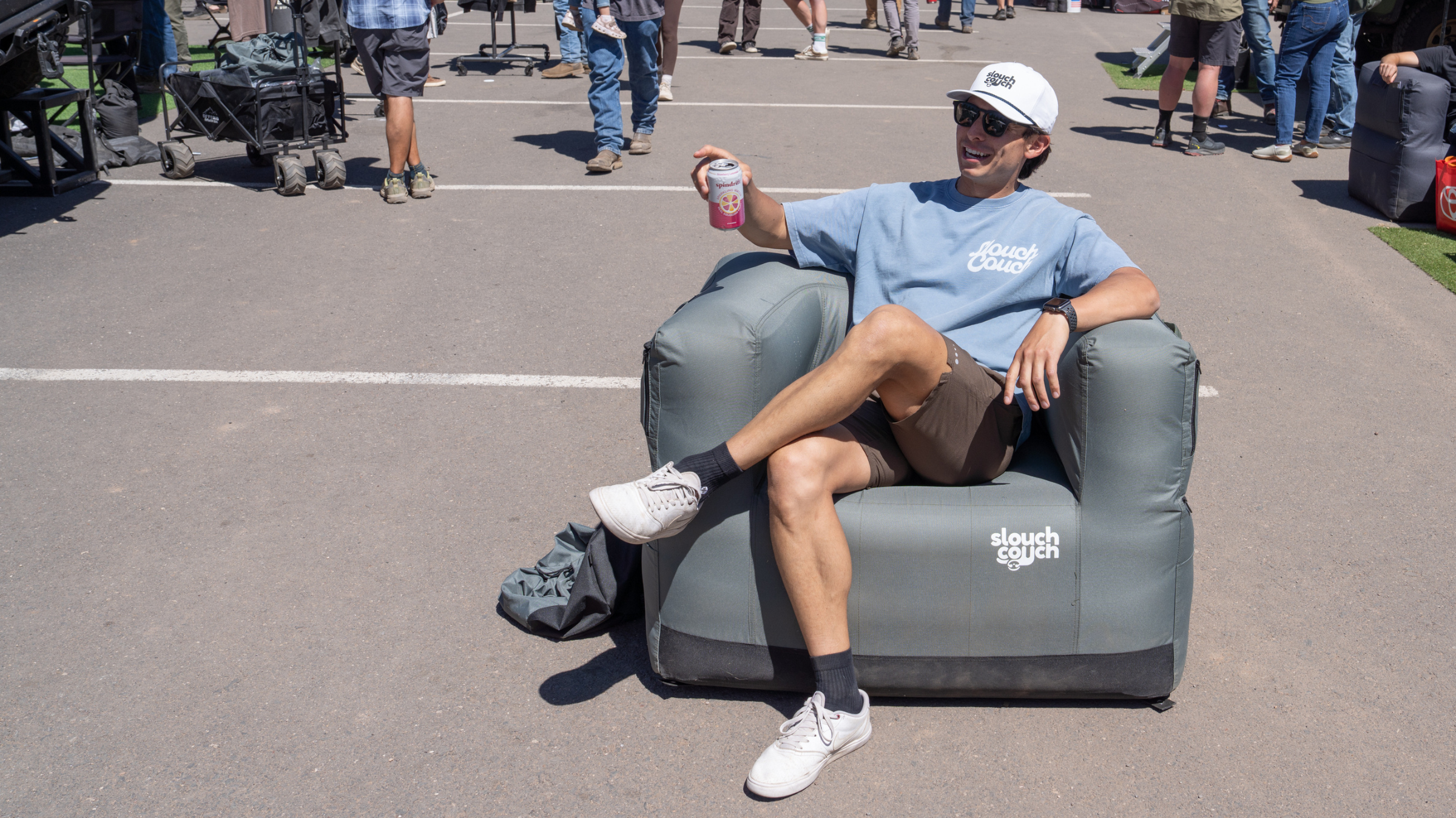 man in blue T-shirt, hat and shorts holds a Spindrift and sits on gray inflatable chair with Slouch Couch written on it on asphalt at Overland Expo West