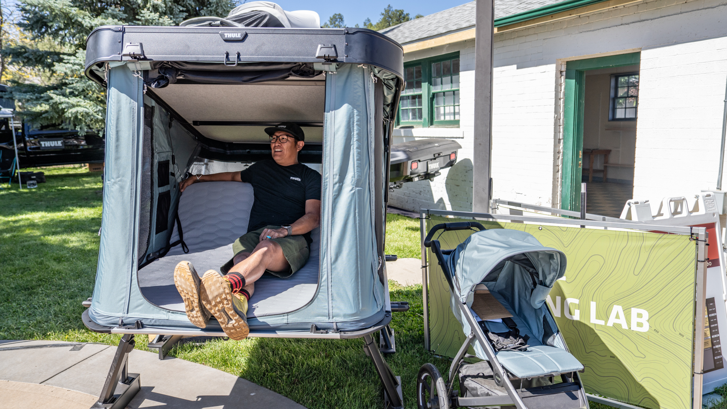 Man sitting in light blue version of Thule Widesky rooftop tent set up over grass and sidewalk outside at 2026 Overland Expo West