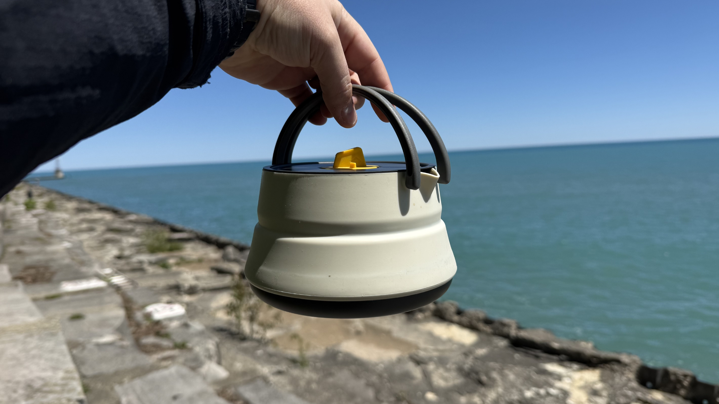 A hand holds up a lightweight Sea to Summit collapsible kettle against a sunny blue sky and vast lake background, illustrating compact gear for camping coffee.