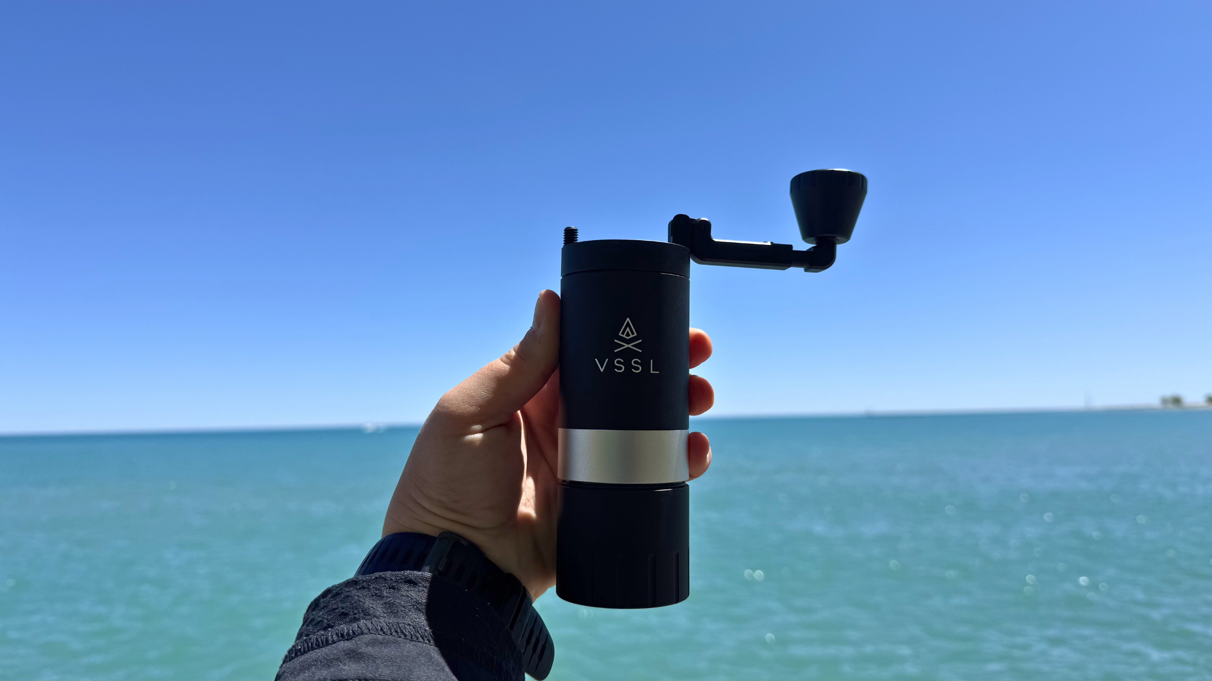 A hand holds a sleek black VSSL Java G25 manual burr coffee grinder up against a clear blue sky and sparkling lake water to prepare fresh camping coffee.