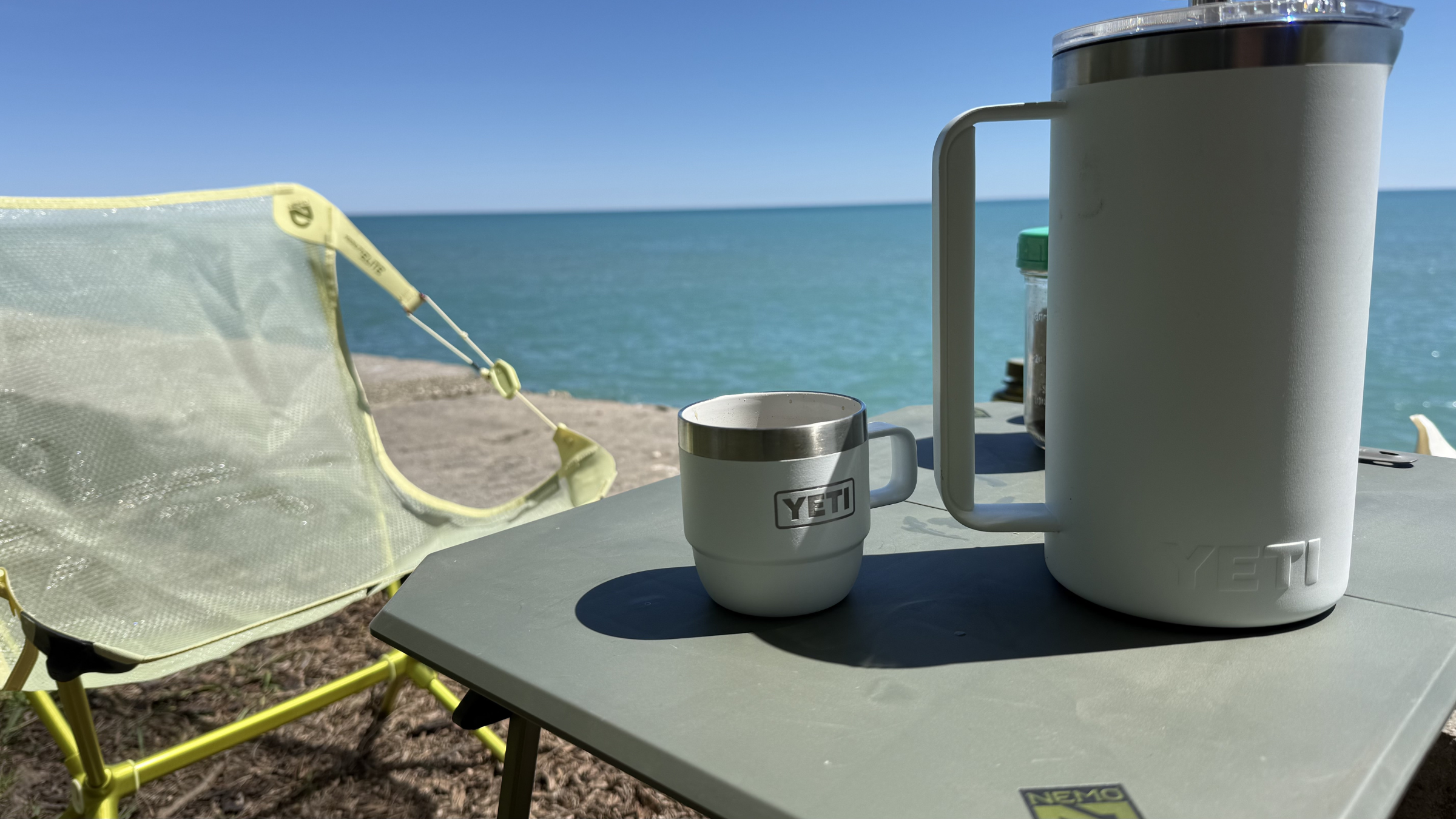 A white YETI stackable mug and a vacuum-insulated YETI French press rest on a camp table overlooking a scenic blue lake, perfect for enjoying hot camp coffee.