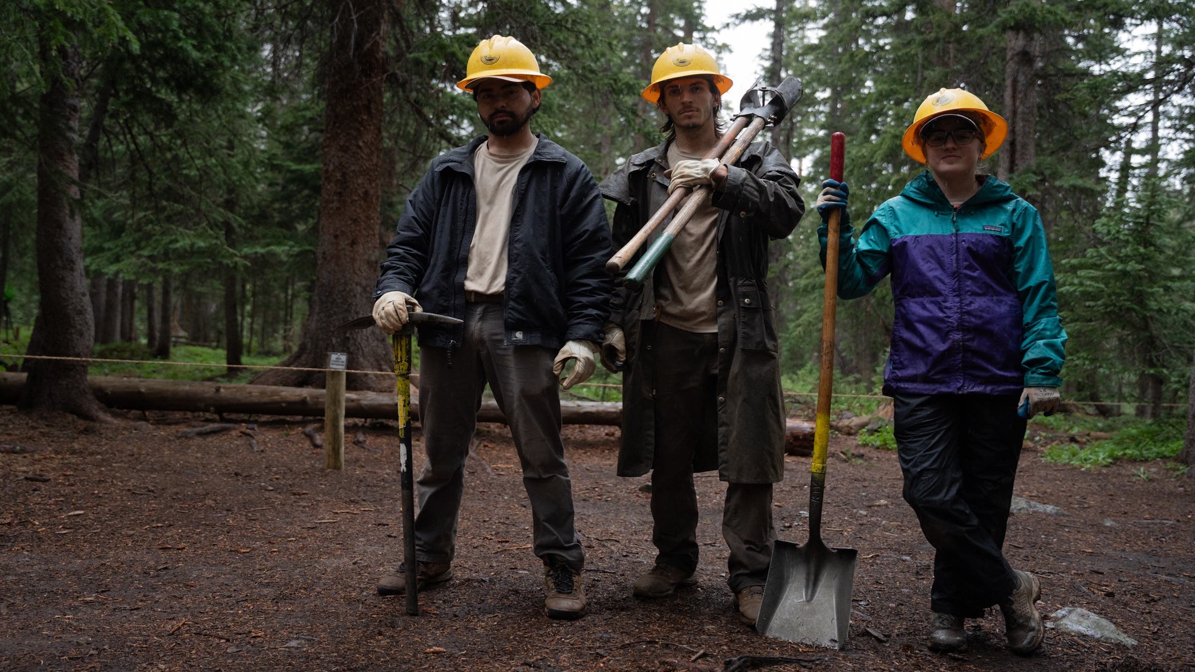 A trio of three crew workers stand with shovels and yellow hard hats on the trail