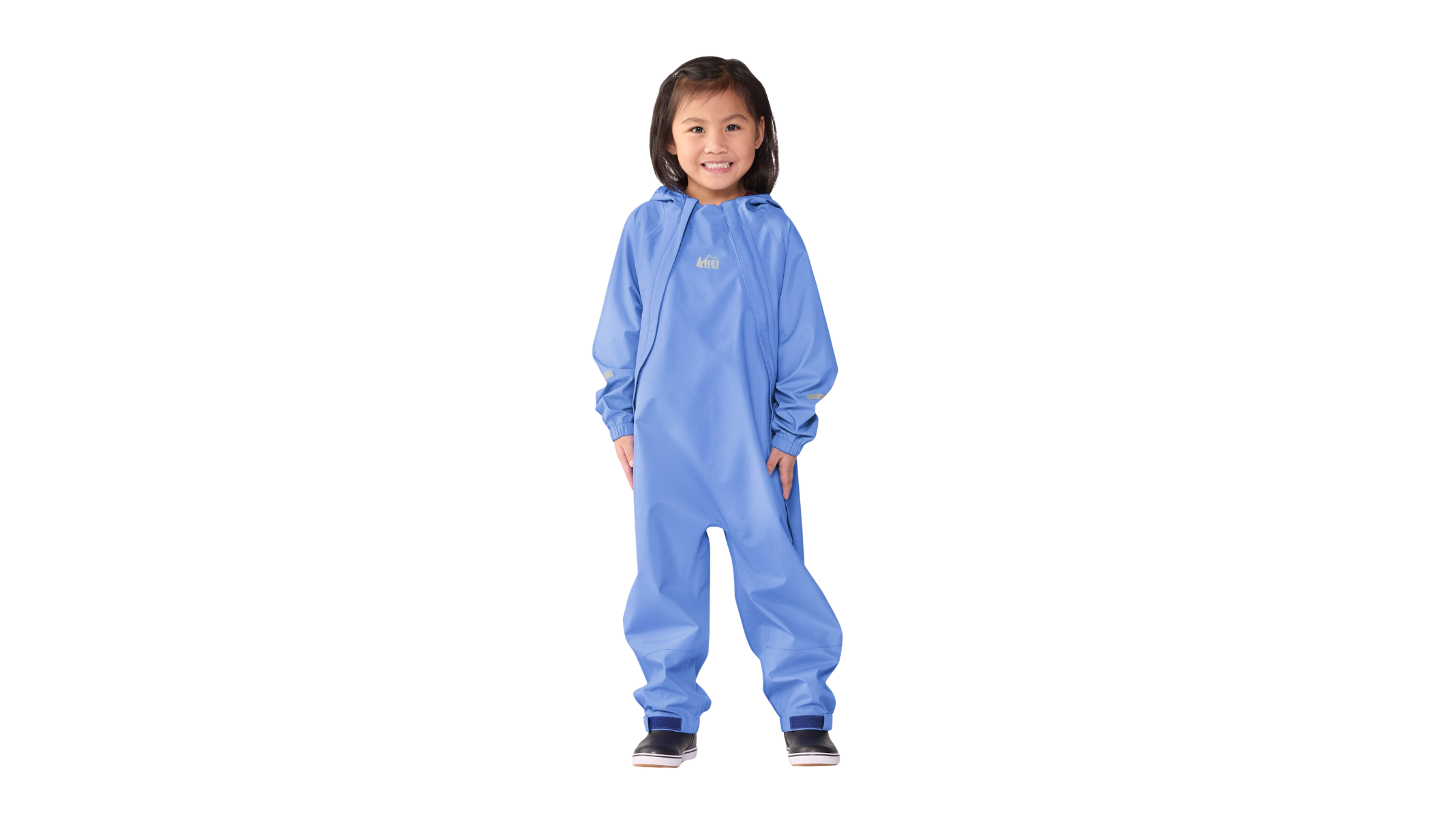 A smiling young girl wearing a full-body, light blue waterproof REI rain suit, a crucial foul-weather outerwear piece for any summer camp packing list.