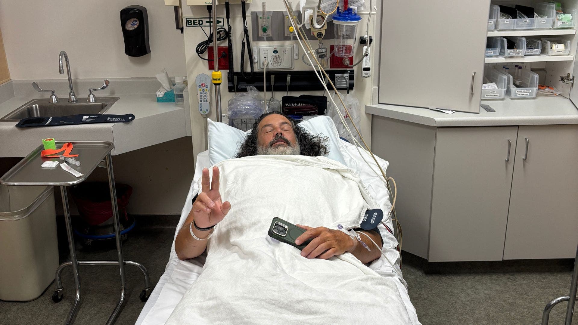 matthew wappett showing in a hospital bed hooked up to IVs, holding.a peace sign