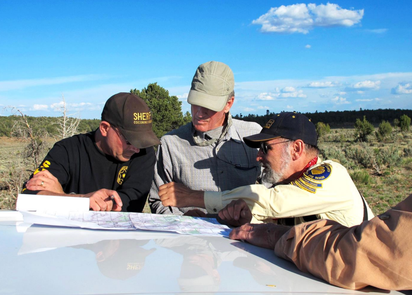three men are shown looking at a map on the hood of a vehicle during the search