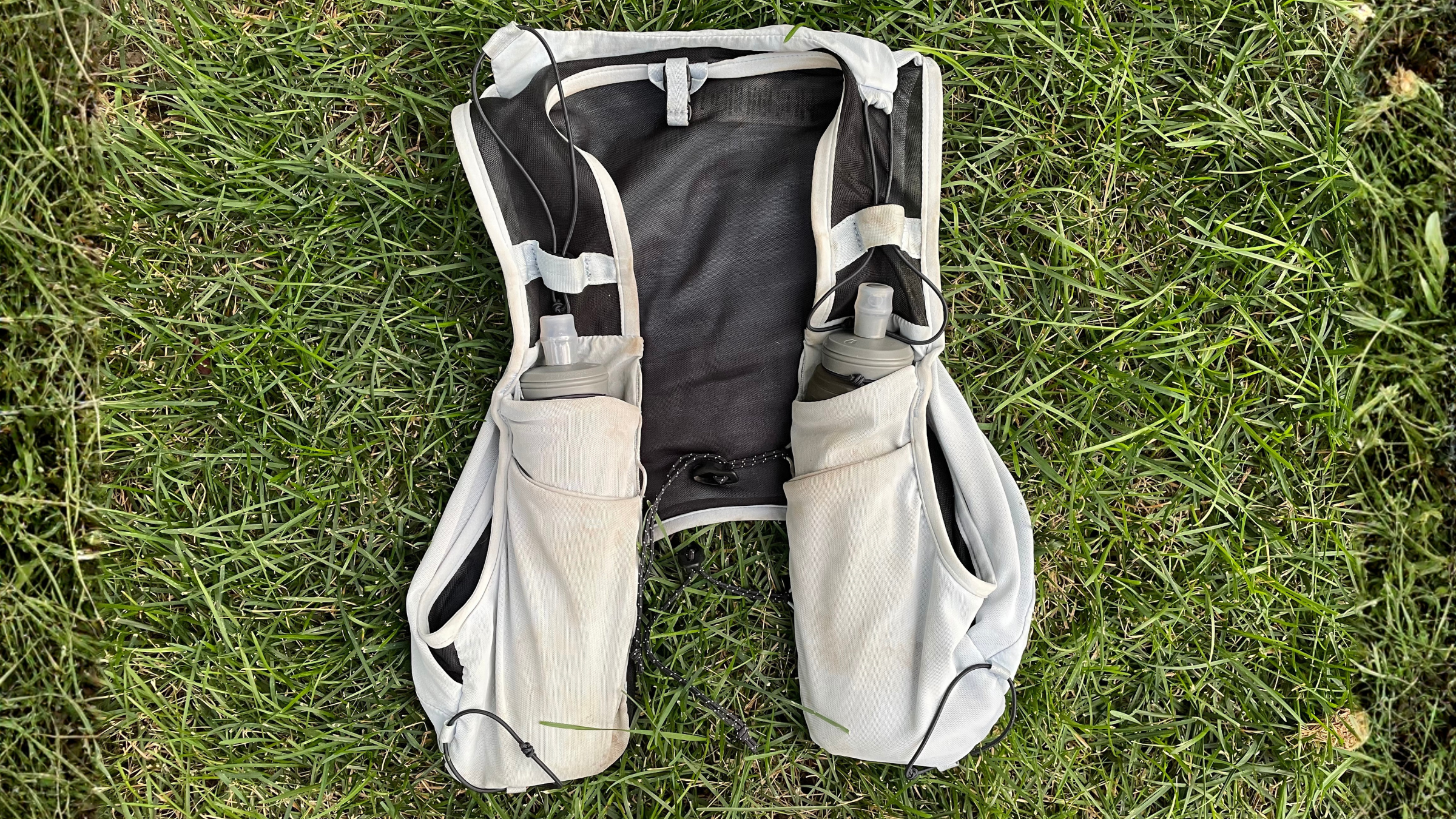 Light grey trail running hydration vest with dual soft flasks tucked into the front pockets, resting flat on green grass.