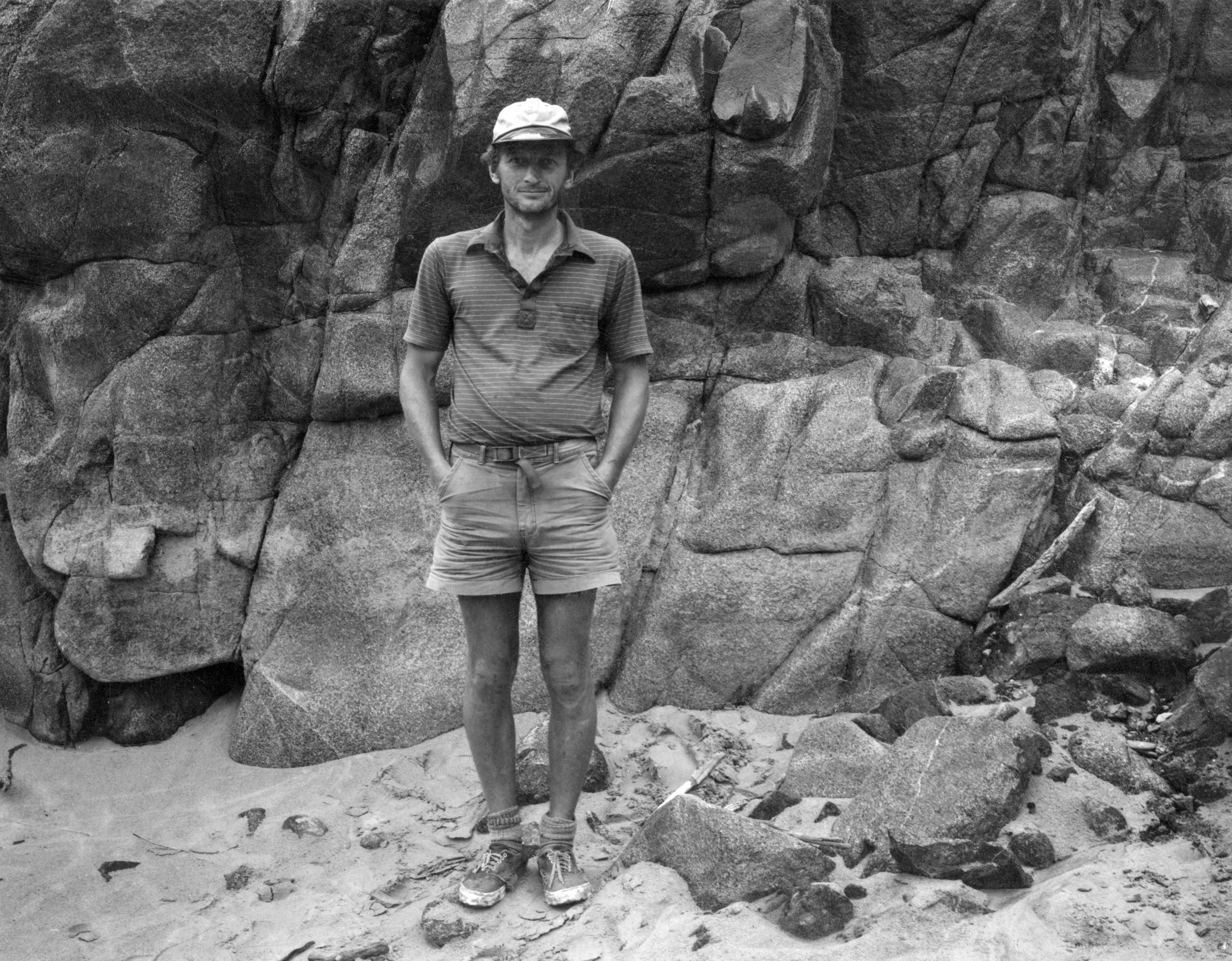 black and white photograph shows bill ott standing against a rock wall wearing shorts and sandals