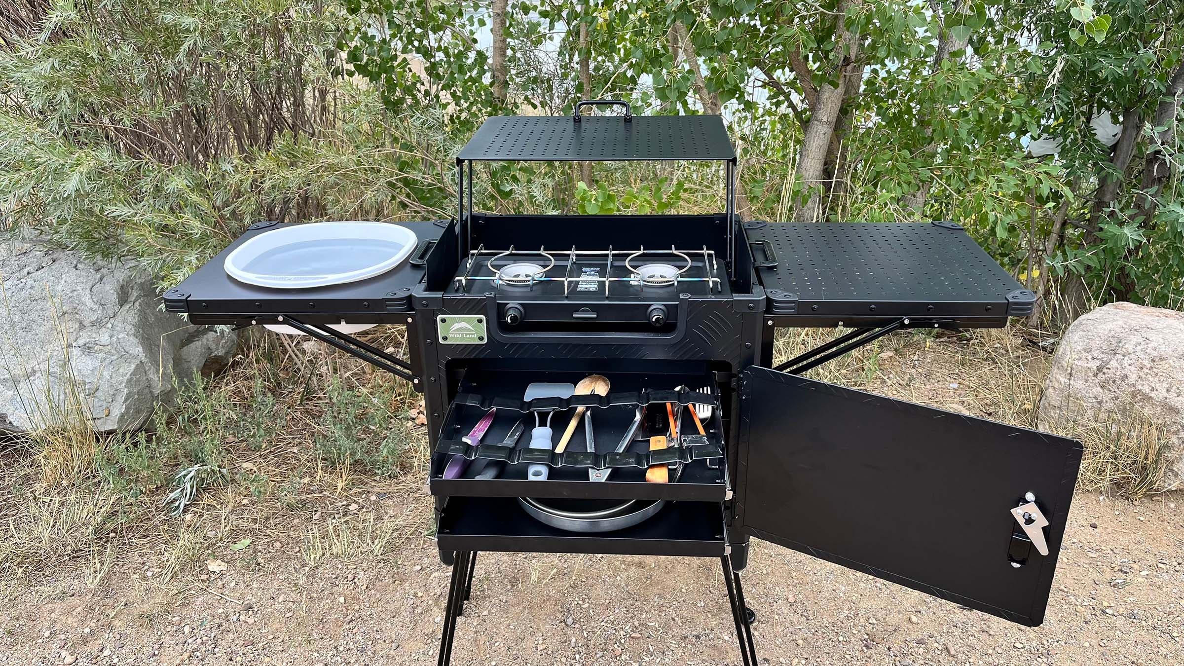 The Cinch Kitchen Cruiser fully set up with a collapsible sink on one side table, the two-burner stove in the center, and a drawer open below revealing stored cookware and utensils.