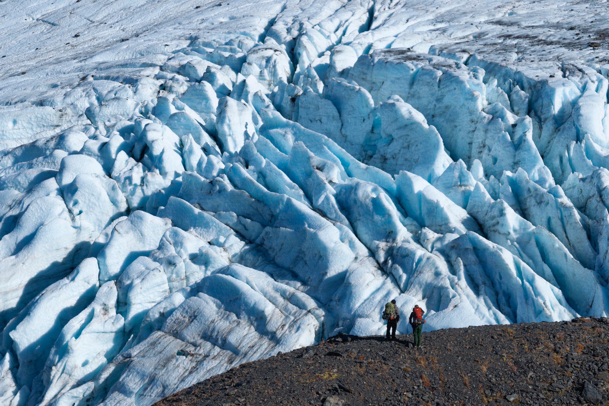 two hikers shown standing below raven glacier along crow pass