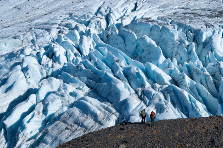 two hikers shown standing below raven glacier along crow pass