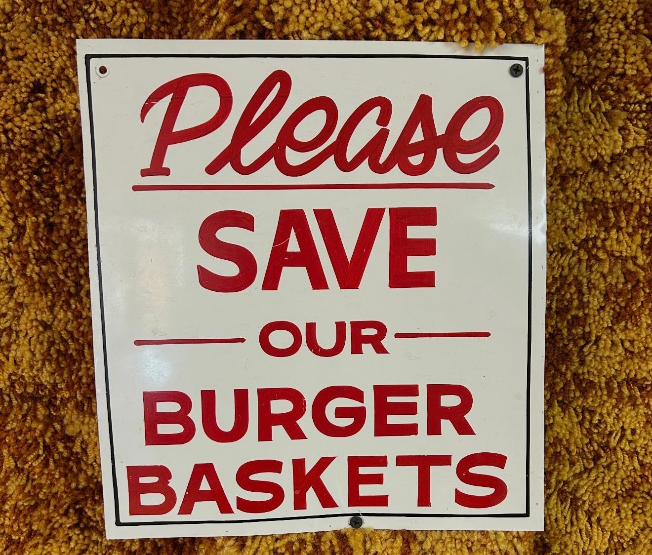 A sign reading "please save our burger baskets" shown hanging against a yellow shag carpet wall