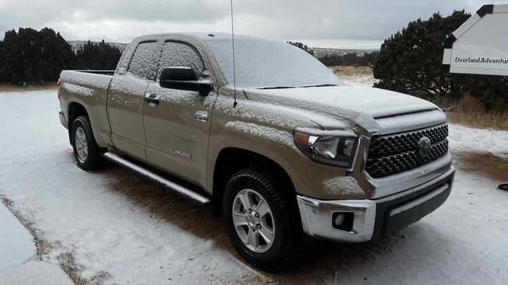 Tan Toyota Tundra covered in fresh snow, parked outside a garage next to an overland trailer.